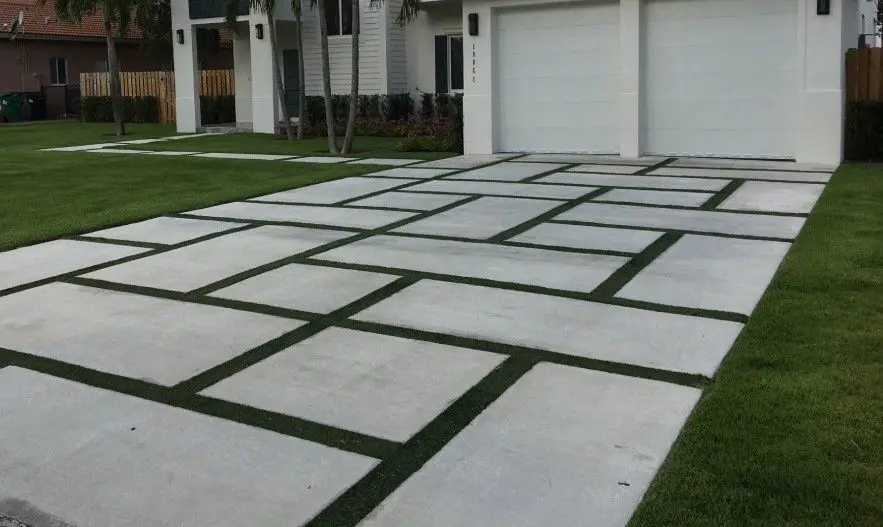 A modern driveway with a geometric design features large rectangular and square concrete slabs separated by strips of Pet Turf. The driveway, inspired by Palm Springs aesthetics, leads to a double garage with white doors, attached to a contemporary house. The lawn is well-maintained.