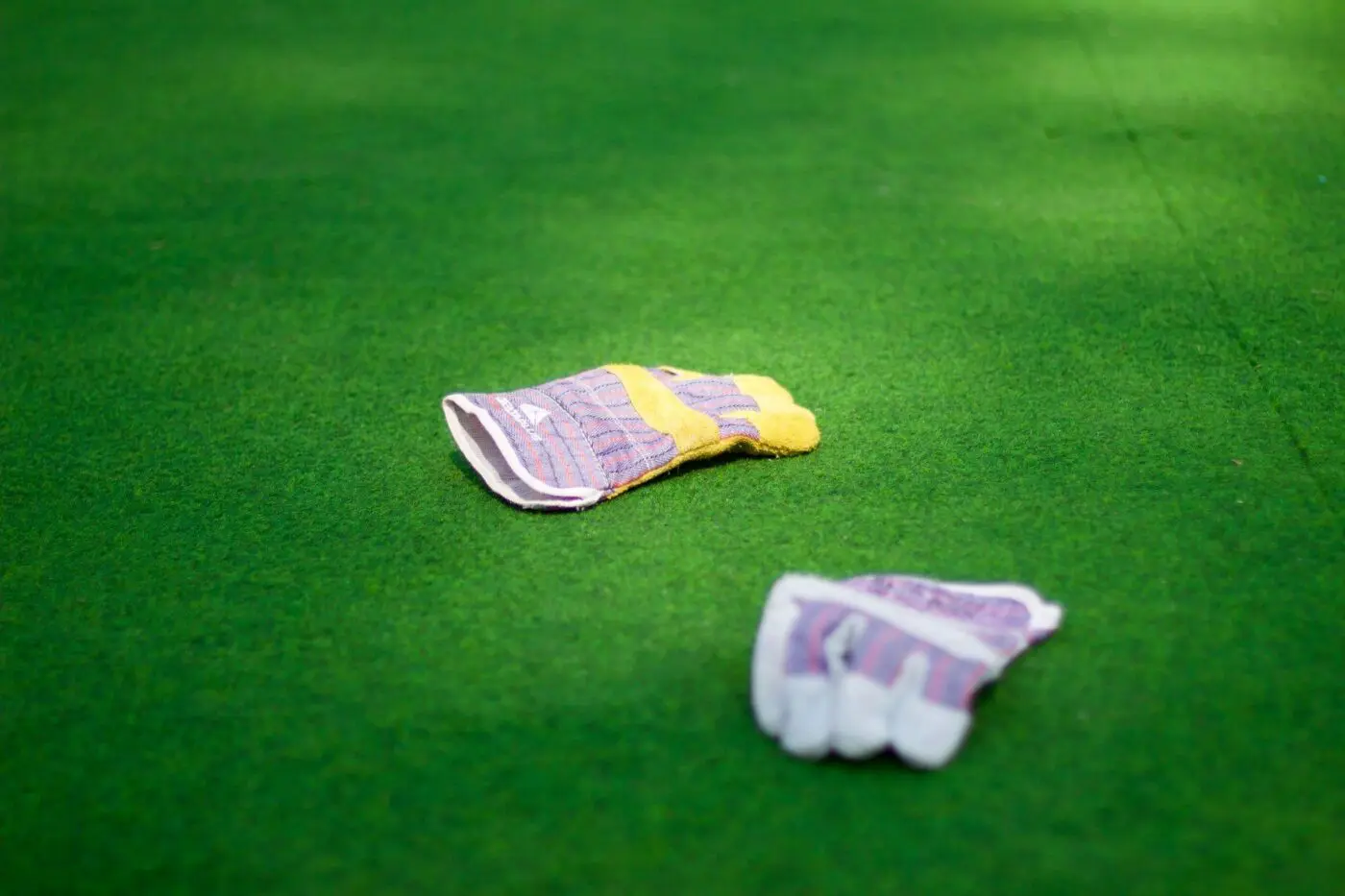 Two gardening gloves lie on a vibrant green lawn reminiscent of a well-maintained sports field astroturf. One glove is slightly closer to the foreground, with both gloves showing a mix of purple, yellow, and white colors. The scene is brightly lit, highlighting the gloves against the lush grass background.