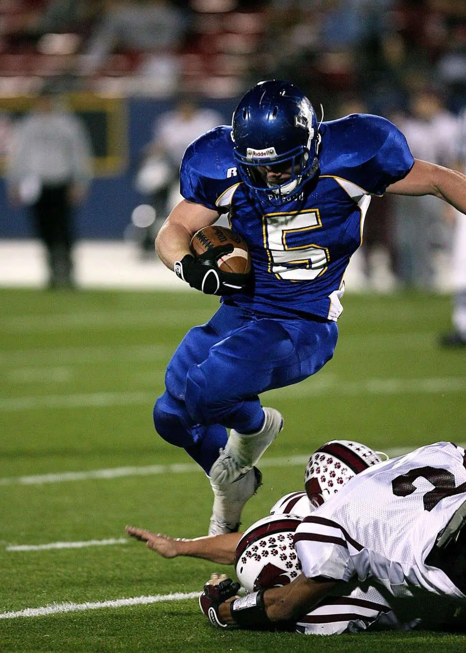 A football player wearing a blue uniform with the number 5 runs with the ball, avoiding a tackle from two defenders in white uniforms on Coachella Valley Turf. The action unfolds on this premier sports field astroturf installer’s site, with a crowd blurred in the background.