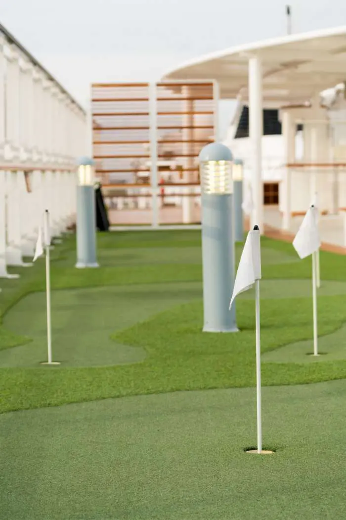 A Palm Springs-inspired mini-golf course on a ship deck features holes marked by white flag poles. The green artificial turf, installed by a professional Artificial Turf Installer, complements ship railings and deck chairs in the background. Two lantern-style lights add charm to the scene.