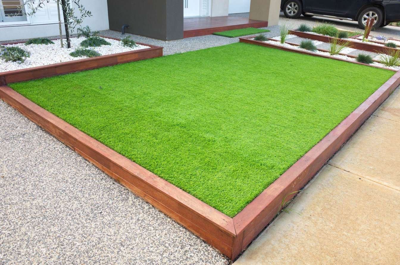 A neatly trimmed front yard in the Palm Springs style features a rectangular patch of vibrant green artificial grass edged with wooden borders. Surrounding areas have white gravel with sparse, neatly arranged plants, contributing to a modern and minimalist landscape design by Coachella Valley Turf.